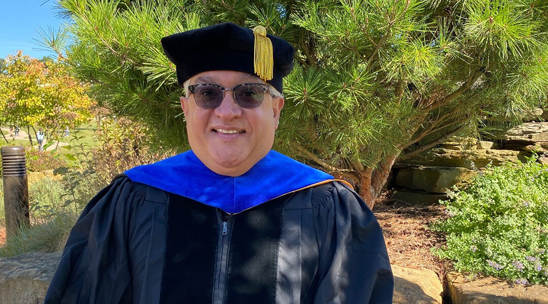 Alum Juan Del Valle stands outside on a sunny day in his doctoral regalia.