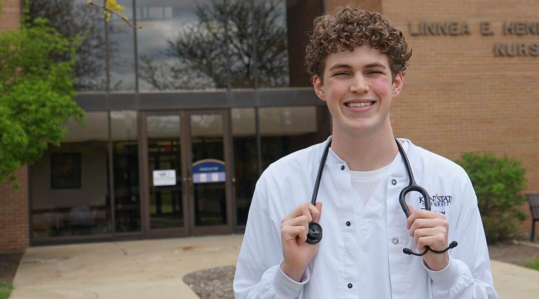 Caleb Faust, wearing his Kent State University College of Nursing scrubs, smiles outside of Henderson Hall.