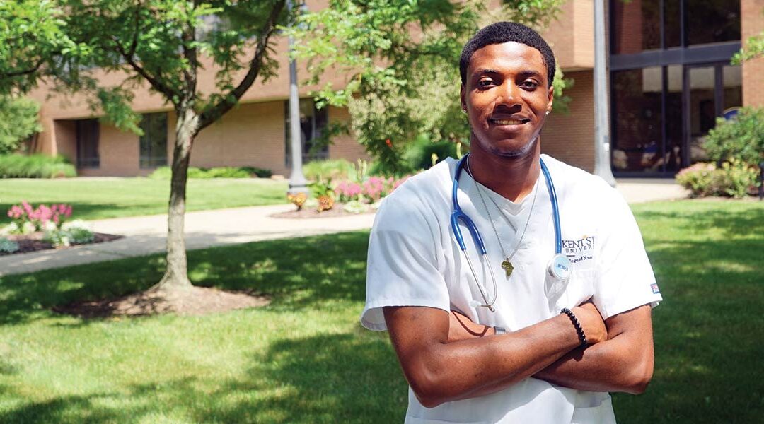 Chukwuemeka (Emeka) Chukwukere wearing his Kent State University College of Nursing scrubs, stands in front on Henderson Hall on the Kent campus on a sunny day with his arms crossed.