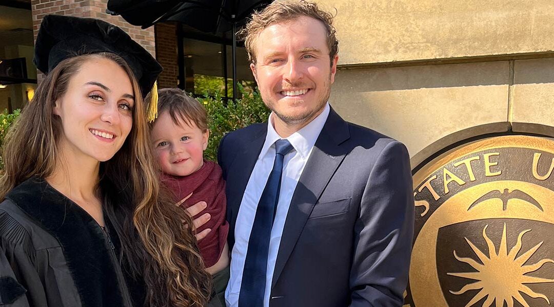 Jasmine Bonder (left) in graduation regalia with her husband, Adam (right) who is holding their young son.