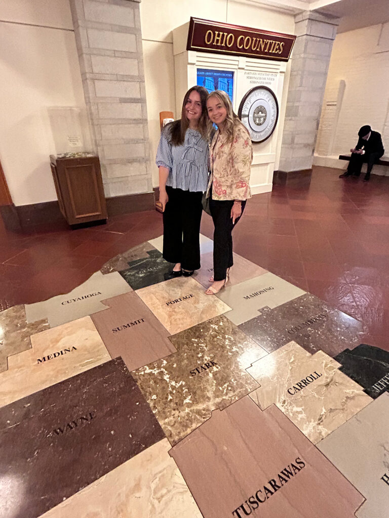 A large graphic map of Ohio's counties is on the floor. Lily Baker (left) and Alanna Lawson (right) stand at the top right corner of Portage county for a photo.  